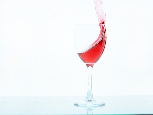 Vinho tinto derramando o movimento na taça de vidro com fundo de vidro branco. salpique com uma gota de vinho tinto ou um saboroso líquido vermelho. respingos de vinho. um vidro fica em uma superfície branca brilhante e isolado no branco.