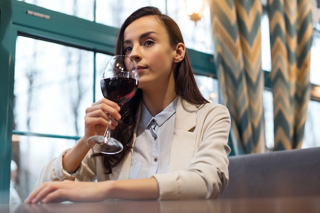 Vinho tinto. mulher pensativa, simpática e melancólica, levantando a taça de vinho enquanto está sentada perto da janela e aproveitando o tempo