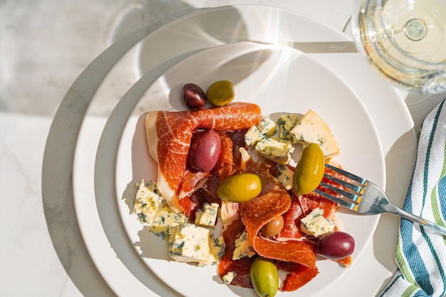 Foto vino blanco fresco en un vaso cerca de un plato con lonchas de jamón, queso paloma y varias aceitunas