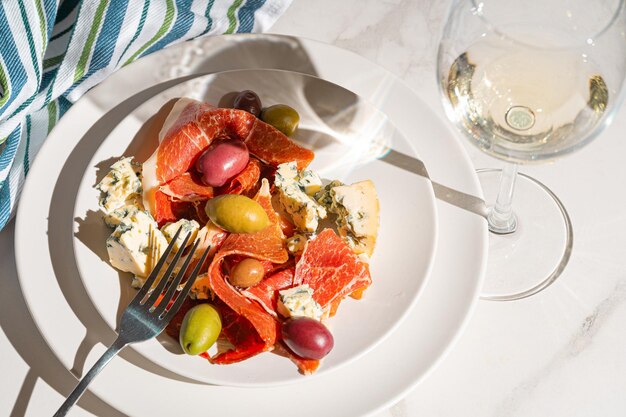 Foto vino blanco fresco en un vaso cerca de un plato con lonchas de jamón, queso paloma y varias aceitunas