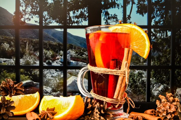 Vino caliente Bebida navideña caliente con especias en el alféizar de una ventana, junto a la ventana. Rodajas de naranja, ramitas de canela, piñas. Ambiente de vacaciones, estilo rústico. La idea de crear tarjetas de felicitación.