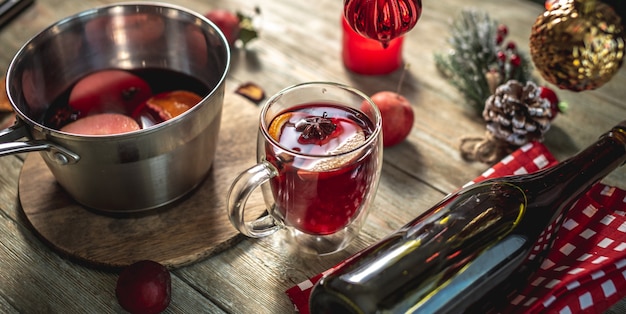 Vino caliente caliente aromático picante en una cacerola y un vaso transparente sobre una mesa de madera con accesorios festivos. Concepto de un ambiente de vacaciones acogedor, año nuevo y humor navideño.