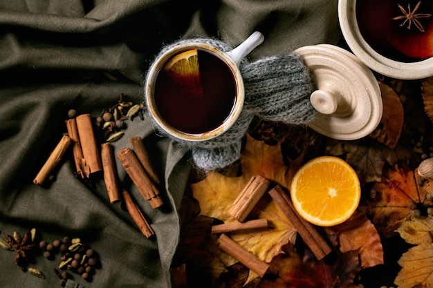 Vino caliente caliente en olla de cerámica y taza en bufanda con especias, naranja y hojas de otoño sobre un mantel de lino oscuro. Endecha plana. Ambiente cálido y acogedor. Endecha plana