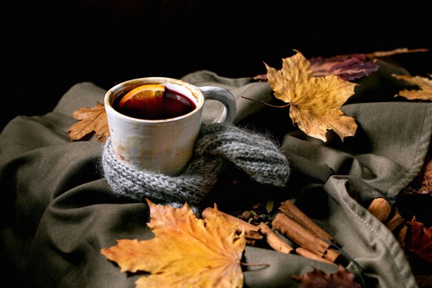 Vino caliente caliente en taza de cerámica en bufanda con especias, naranja y hojas de otoño sobre un mantel de lino oscuro. Bebidas alcohólicas calientes y acogedoras. Copia espacio