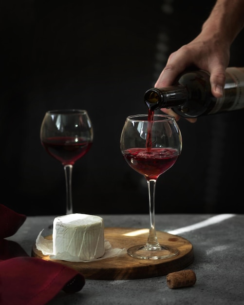 El vino tinto se vierte en un vaso de vino y queso en la mesa Manos masculinas queso brie y vino