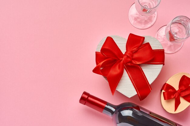 Vino, vasos y caja de regalo en forma de corazón con una cinta roja sobre fondo rosa. Postal del concepto del día de San Valentín. Vista superior.