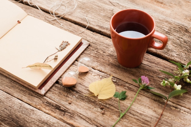 Vintage Album mit Herbariumpflanzen und Sanduhr mit Tasse Tee auf alter Holzoberfläche