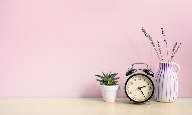 Vintage despertador y planta casera en maceta blanca sobre un escritorio de madera sobre un fondo de pared rosa.