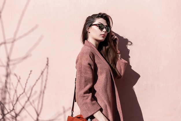 Vintage retrato atractivo joven encantadora en gafas de sol con estilo oscuro con pelo largo en elegante abrigo con bolso cerca de la pared rosa vintage al aire libre. Modelo hermosa hermosa chica disfruta del sol de primavera.