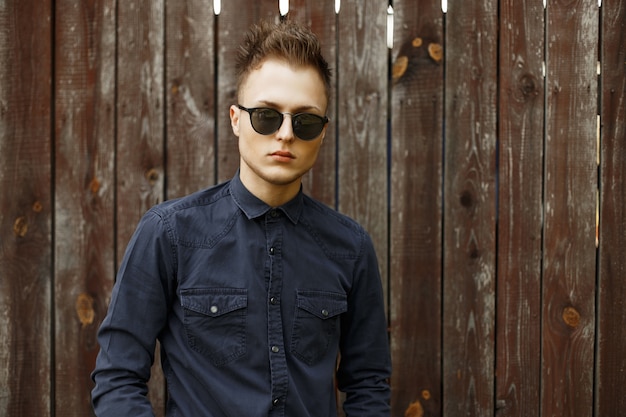 Vintage retrato de un joven con gafas de sol y una camisa azul cerca de una pared de madera vieja