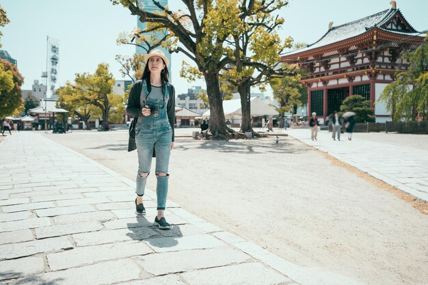 Vintage-Stil der jungen asiatischen Touristenfrau in voller Länge mit Hut und Rucksack, die in der Stadt in der Nähe des Shitennoji-Tempels in Osaka, Japan, herumläuft. Reise- und Tourismuskonzept. Reisender mit Kamera.