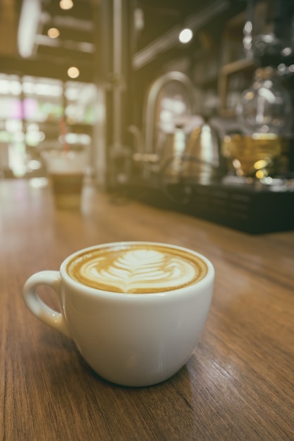 Vintage tono taza de café con leche arte. Café del capuchino en la tabla de madera con el fondo del café de la falta de definición.