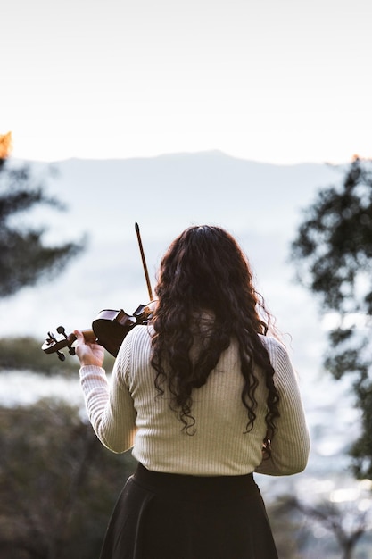 Violinista feminina morena vista de trás tocando violino lá fora na montanha. Vertical