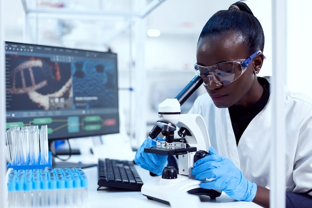 Virologista de etnia africana, sentado em seus tubos de ensaio puros no local de trabalho, olhando através do microscópio. Cientista negro da saúde no laboratório de bioquímica, usando equipamento esterilizado.