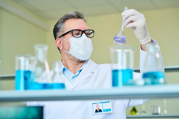 Virólogo masculino maduro con máscara protectora mirando líquido en vaso de precipitados