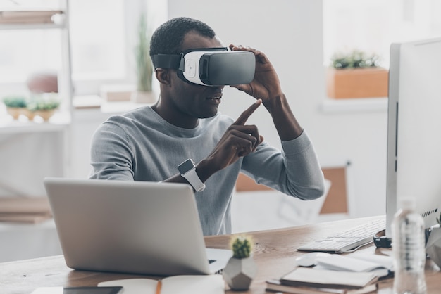 Virtuelle Arbeit. Hübscher junger Afrikaner mit VR-Headset, der in die Luft zeigt, während er am Schreibtisch im Kreativbüro sitzt