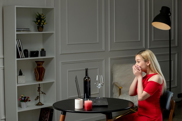 Virtuelle liebe, süßes mädchen in rotem kleid bei ferntermin mit wein und kerzen, die den mund bedecken