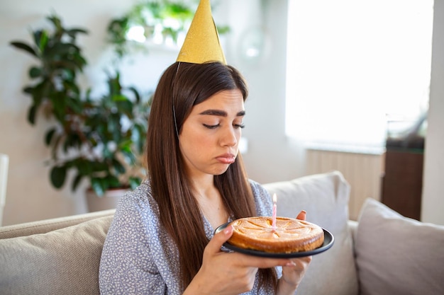 Virtuelle Party während der Sperrung Verärgerte und frustrierte alleinstehende afroamerikanische Frau mit Partyhut, die online Geburtstag feiert Dame, die eine Videokonferenz mit Kuchen und Kerze auf dem Boden hält