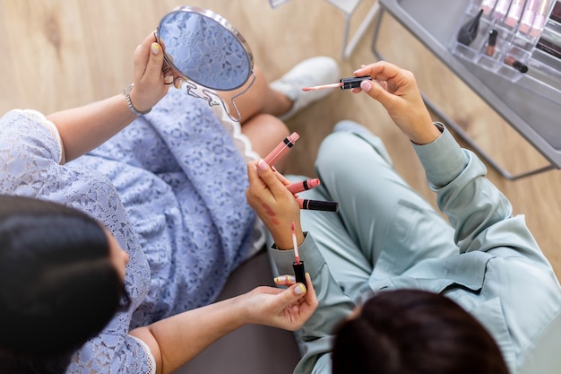 Visagista feminina, vista de cima, ensinando uma cliente aplicando brilho labial na aula de maquiagem profissional