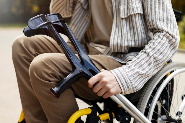 Visão aproximada de muletas nas mãos de uma pessoa Um homem se senta em uma cadeira de rodas segurando muletas na rua