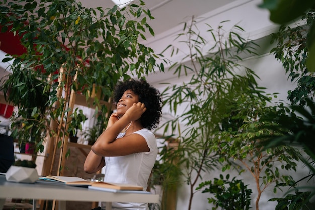 Visão de baixo ângulo de uma jovem afro-americana encaracolada positiva ouvindo música em fones de ouvido sentada na mesa com laptop e sorrindo olhando para longe