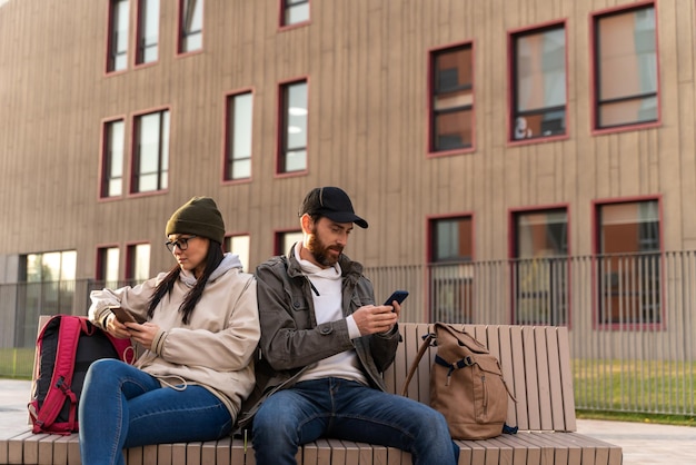 Visão de corpo inteiro do homem chato e da mulher usando seus telefones enquanto estão sentados no banco durante o dia. Cônjuges não se comunicam um com o outro