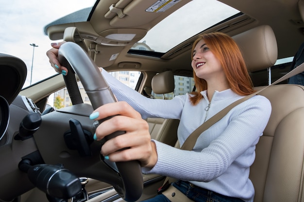 Visão de grande angular do motorista jovem ruiva prendida pelo cinto de segurança, dirigindo um carro sorrindo alegremente.