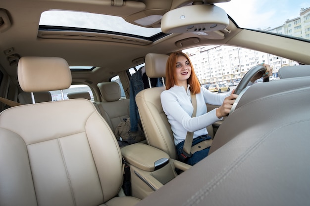 Visão de grande angular do motorista mulher jovem ruiva prendida pelo cinto de segurança, dirigindo um carro sorrindo alegremente.