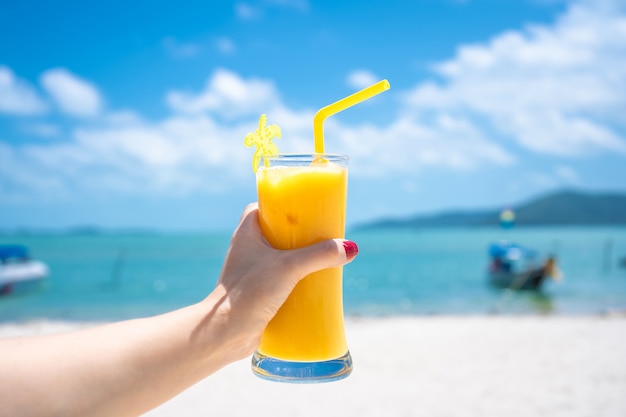 Visão em primeira pessoa. Garota mantém um copo de manga fria de vidro fresco no fundo de uma praia tropical. Areia branca e um barco. Férias de conto de fadas na Tailândia