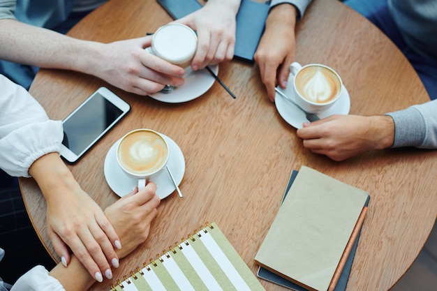 Visão geral das mãos de estudantes universitários com gadgets e bebidas reunidos à mesa no café para uma xícara de cappuccino e bate-papo