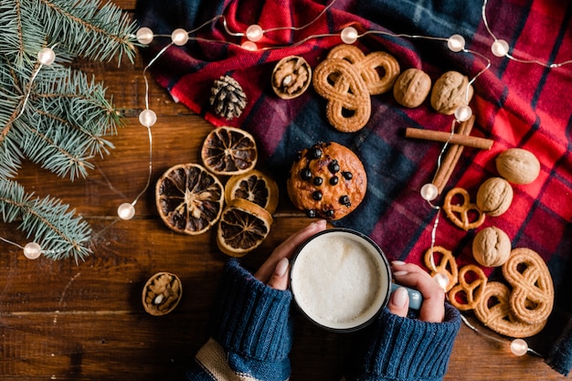 Visão geral das mãos de mulheres em uma camisola de malha segurando uma caneca com uma bebida quente cercada por símbolos e comida tradicional de Natal