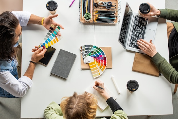 Foto visão geral de três jovens designers criativos sentados à mesa e escolhendo cores para o novo projeto na paleta, enquanto um deles faz anotações