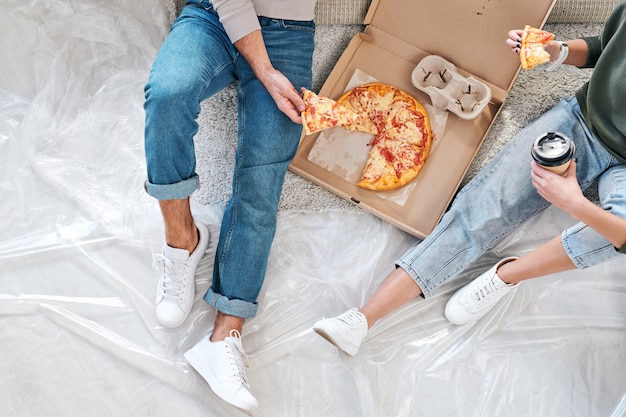 Visão geral de um jovem casal em trajes casuais, sentado no chão e comendo pizza na caixa e café após a mudança para um novo apartamento ou casa