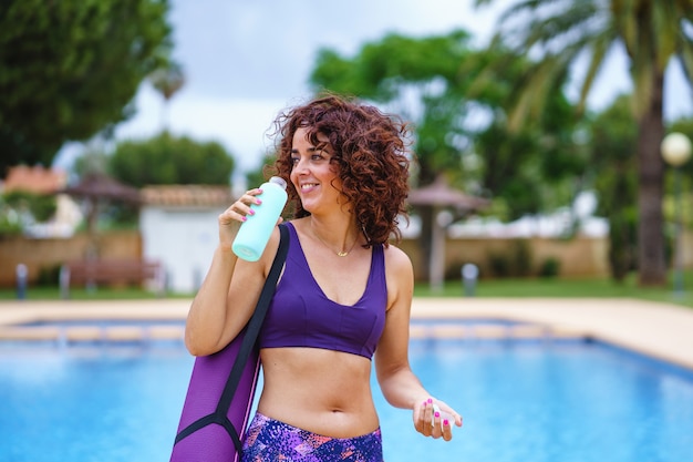 visão horizontal de mulher de cabelo encaracolado vestida com uma roupa de ioga, segurando água potável. Estilo de vida de hidratação e fitness. Exercício e hábitos esportivos saudáveis no verão.