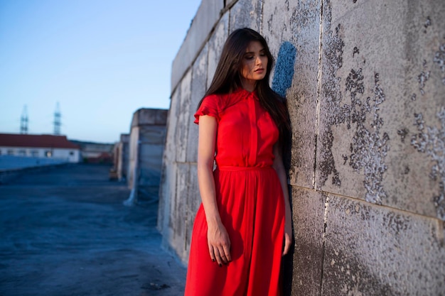 Visão horizontal jovem linda de vestido vermelho com maquiagem posando no telhado no fundo do céu limpo por do sol