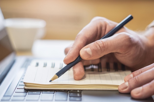 Visão nítida da mão de um homem transcrevendo em um bloco de notas em um laptop de última geração com um pano de fundo desfocado