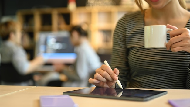 Visão recortada da jovem criativa segurando a xícara de café e usando tablet digital em seu local de trabalho