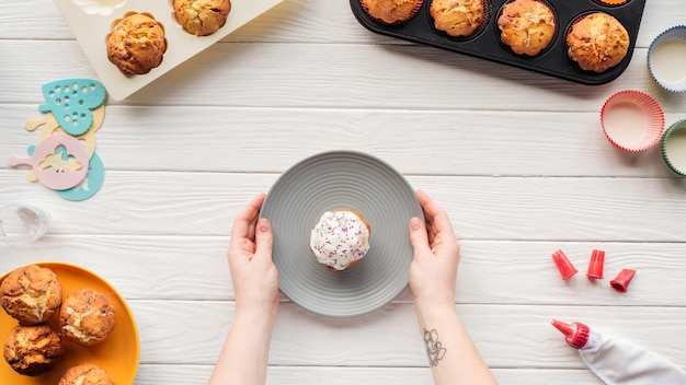 Foto visão recortada da mulher segurando o prato com cupcake na mesa com bandejas e ferramentas de cozimento