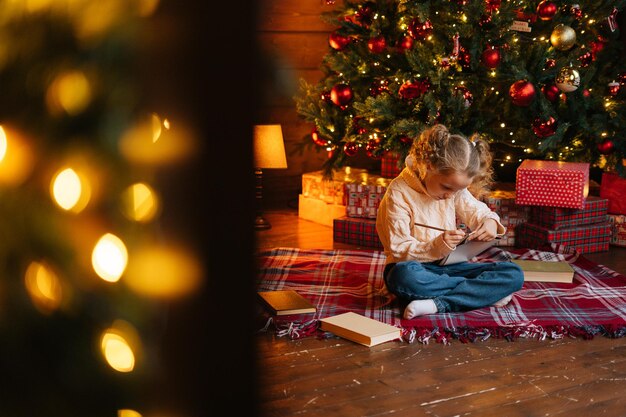 Visão remota de uma garotinha loira e encaracolada, focada, escrevendo carta para o papai noel sentado