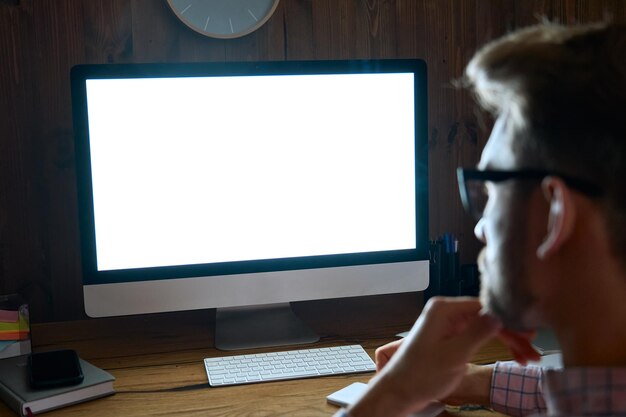 Visão sobre o ombro de um homem de negócios usando um computador de mesa olhando para um monitor de tela branca para publicidade trabalhando até tarde e aprendendo on-line no site assistindo a um webinar no escritório em casa