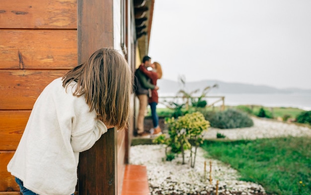 Visão traseira da garotinha espionando casal apaixonado se beijando ao lado de uma casa de madeira