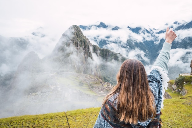 Visão traseira da mulher caucasiana levantando o braço e olhando para Machu Picchu usando o poncho peruano