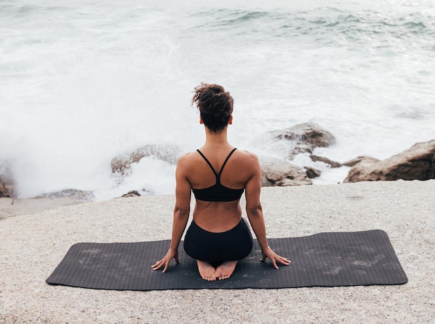 Visão traseira da mulher magra em roupas esportivas sentada no tapete olhando para o oceano