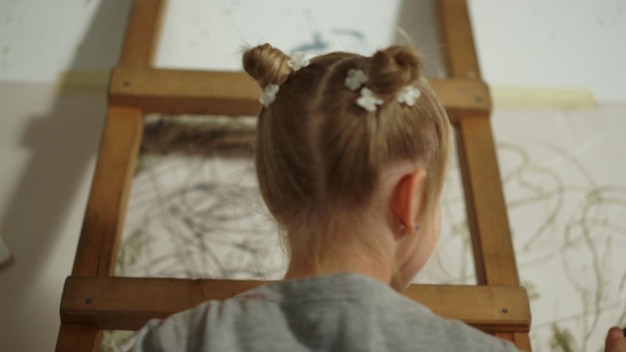 Visão traseira da pequena senhora criando obras de arte abstratas na parede branca no estúdio de arte Menina talentosa desenhando em tela dentro de casa Jovem pintora em pé na escada dentro