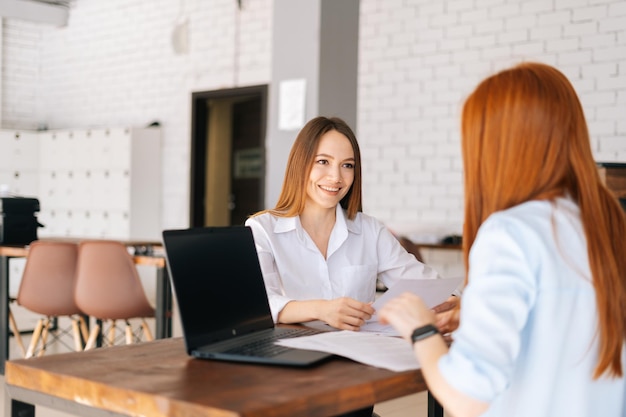 Visão traseira de duas mulheres de negócios jovens positivas discutindo novas ideias de compartilhamento de projetos sentados na mesa em frente um do outro na sala de reuniões do escritório Conceito de coaching e mentoring de negócios