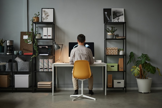 Visão traseira de grande angular para o homem usando o computador no escritório em casa com espaço de cópia do interior de tecnologia moderna