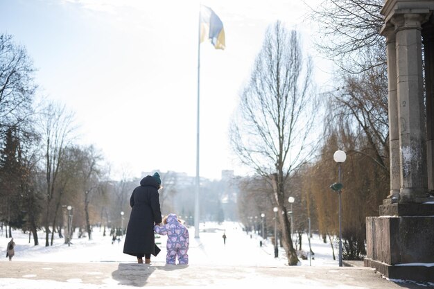 Visão traseira de mãe e filho caminhando em um dia ensolarado e gelado de inverno no parque contra o fundo de um mastro de bandeira com a bandeira ucraniana