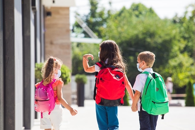 Visão traseira de três alunos que voltam à escola após a pandemia por crianças com máscaras protetoras e mochilas para segurança