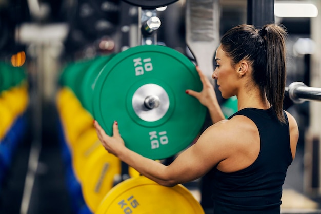 Foto visão traseira de uma forte fisiculturista feminina colocando placas de peso em uma barra em uma academia