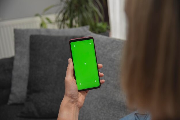 Visão traseira de uma jovem europeia que está usando a tela do smartphone chroma keyEla está sentada no sofá em sua aconchegante sala de estar